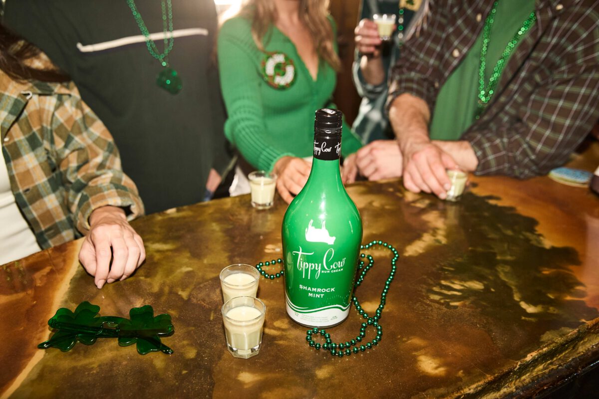 A green bottle of Tippy Cow Shamrock Mint Rum Cream sits on a rustic wooden bar alongside two small poured shots, green beads, and shamrock-shaped glasses. In the background, four friends wearing green St. Patrick's Day clothes and beads raise their glasses in a toast.