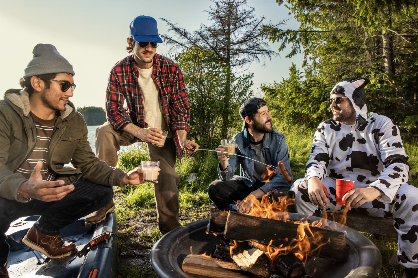 Four friends sitting and standing around a campfire by a lake at sunset. One man wears a cow-patterned onesie while holding a red cup, and another roasts a hot dog over the flames as they all hold glasses of Tippy Cow rum cream.