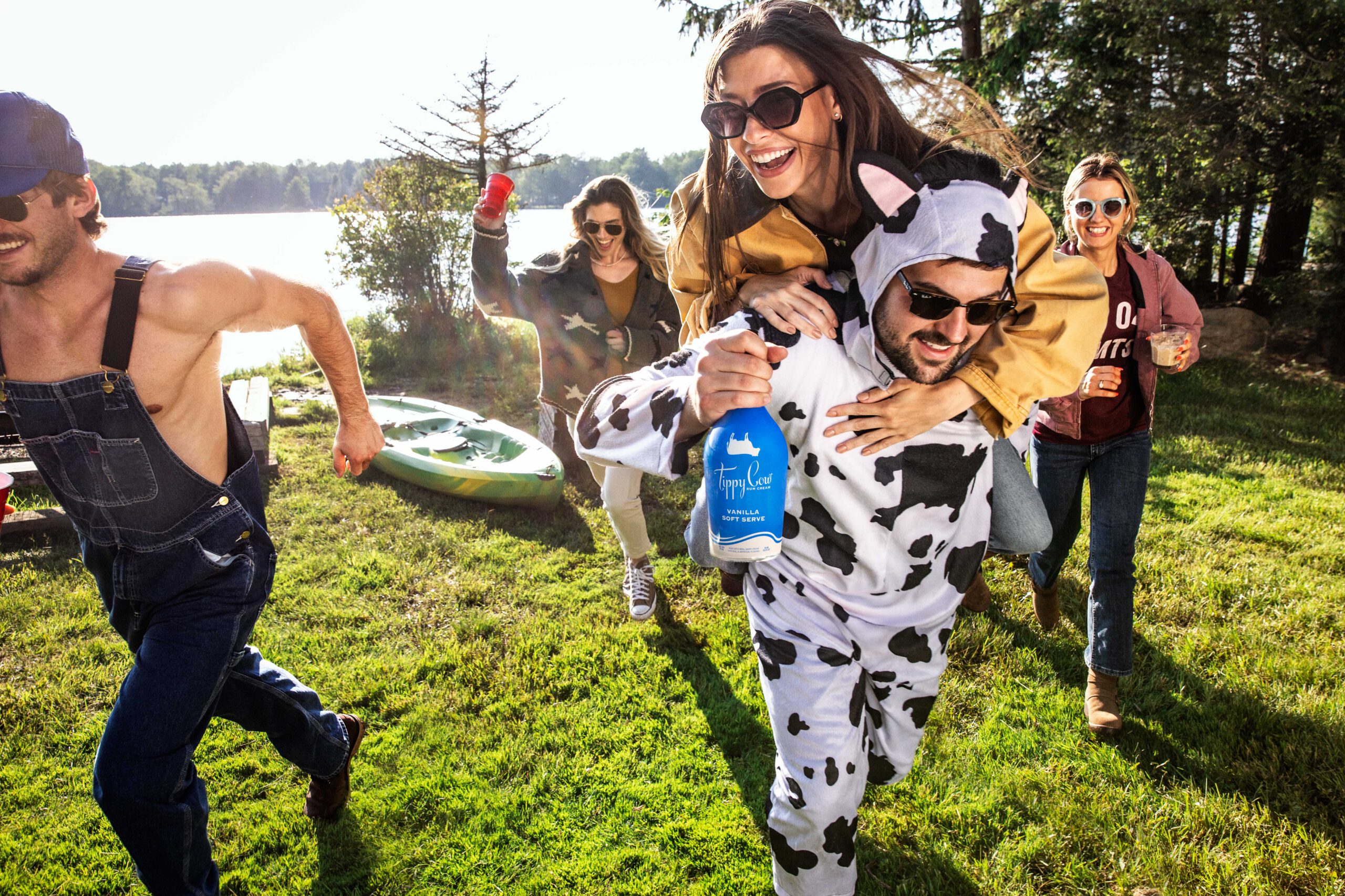 A group of friends laughing and running through a grassy field toward a lake. One person wears a cow-patterned onesie while giving a piggyback ride to a friend and holding a bottle of Tippy Cow Vanilla Soft Serve.