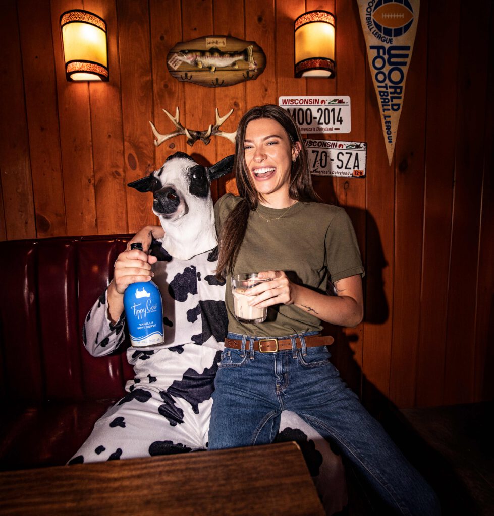 A woman in a green t-shirt laughs while sitting in a red leather bar booth next to a person in a full cow-patterned onesie and a realistic cow mask. The person in the cow suit holds a blue bottle of Tippy Cow Vanilla Soft Serve, and the background features wood-paneled walls with Wisconsin license plates and a deer mount.