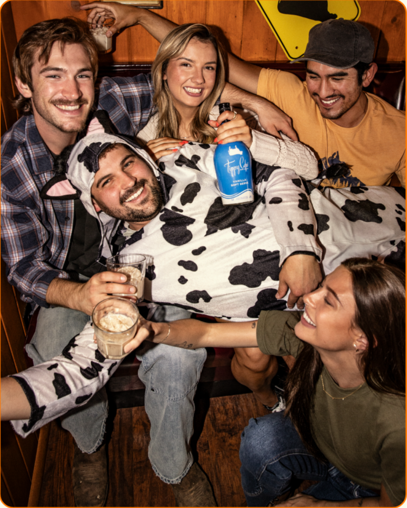 Group of friends laughing in a booth, one person wearing a cow costume while holding blue Tippy Cow vanilla soft serve rum cream bottle.
